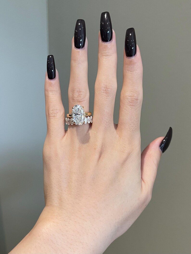 May include: A hand displaying two gold rings. The primary ring features a large, oval-cut diamond. The second ring is a gold band with smaller diamonds. The nails are painted black with a starry design.