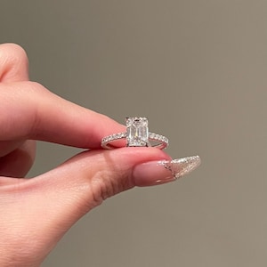 Op de afbeelding: Een zilveren ring met een grote, rechthoekige, smaragd geslepen diamant. De diamant is omgeven door kleinere diamanten die een halo eromheen vormen. De ring is gezet op een dunne band.