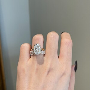 May include: A hand wearing two gold rings. The engagement ring features a large, oval-cut diamond. The wedding band has smaller, round diamonds. The rings are set against a neutral background.