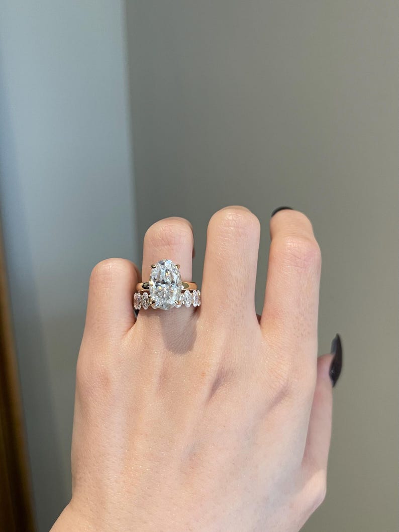 May include: A hand wearing two gold rings. The engagement ring features a large, oval-cut diamond. The wedding band has smaller, round diamonds. The rings are set against a neutral background.