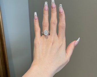 May include: A hand with long, oval-shaped nails with white tips and a delicate design. The hand is wearing a gold band and a large oval-shaped diamond ring. The hand is also wearing a gold bracelet.