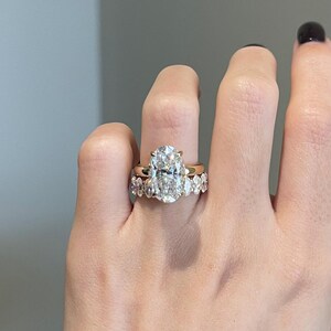 May include: A hand wearing two gold rings. The primary ring features a large, oval-cut diamond. The second ring has a band of smaller, pear-shaped diamonds. The rings are set against a neutral background.