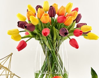 May include: A clear glass vase holding a colourful arrangement of tulips in yellow, orange, red, and purple. The flowers have green stems. The vase is placed on a stack of pink-covered books, with a gold star-shaped ornament nearby.