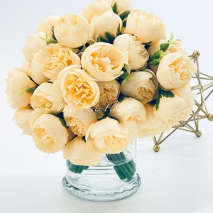 May include: A bouquet of cream-coloured artificial peonies in a clear glass vase. The peonies are arranged in a circular shape and have a realistic appearance.