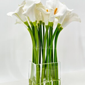 May include: A bouquet of white calla lilies arranged in a clear glass cube vase. The lilies have green stems and are arranged in a symmetrical pattern.