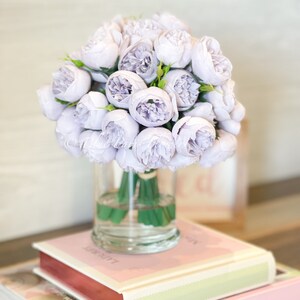 Pu&ograve; includere: Un bouquet di fiori di seta lavanda disposti in un vaso di vetro trasparente. Il vaso &egrave; appoggiato su due libri con copertine rosa.