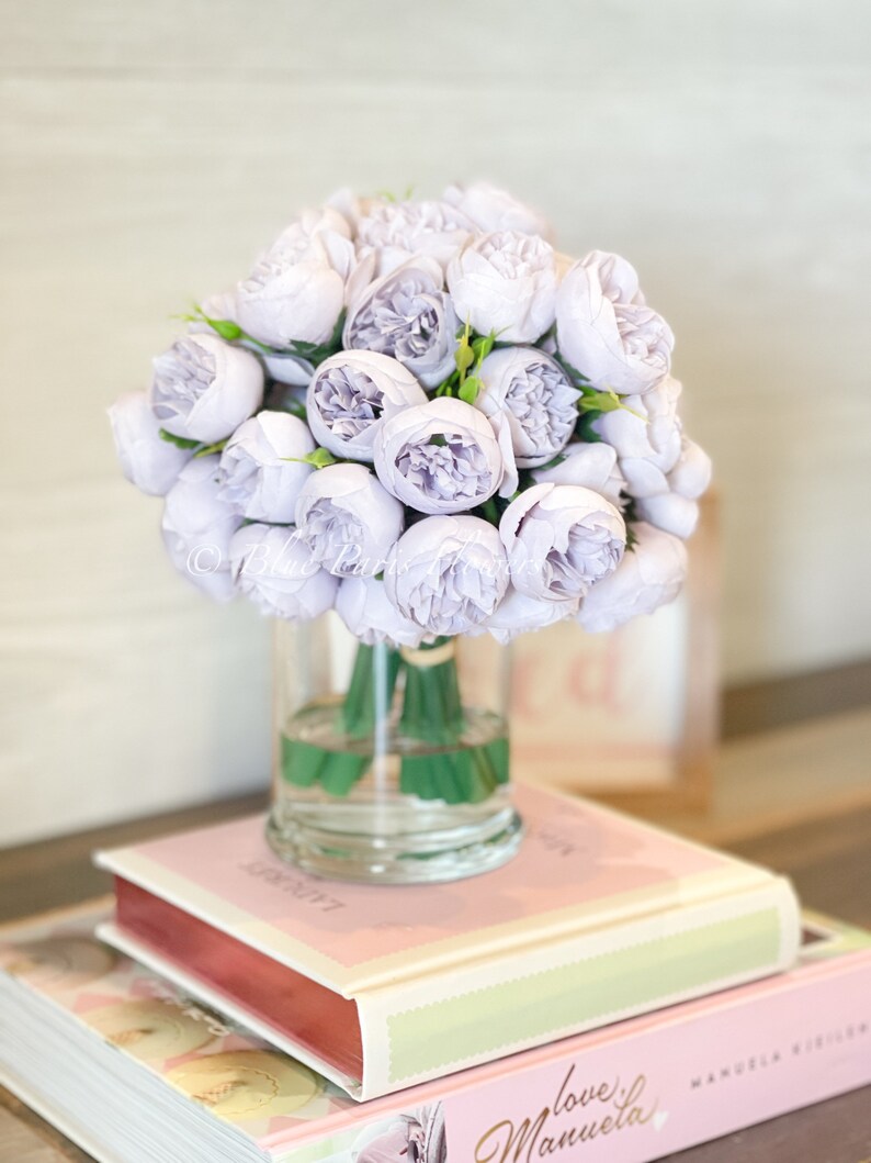 Pu&ograve; includere: Un bouquet di fiori di seta lavanda disposti in un vaso di vetro trasparente. Il vaso &egrave; appoggiato su due libri con copertine rosa.