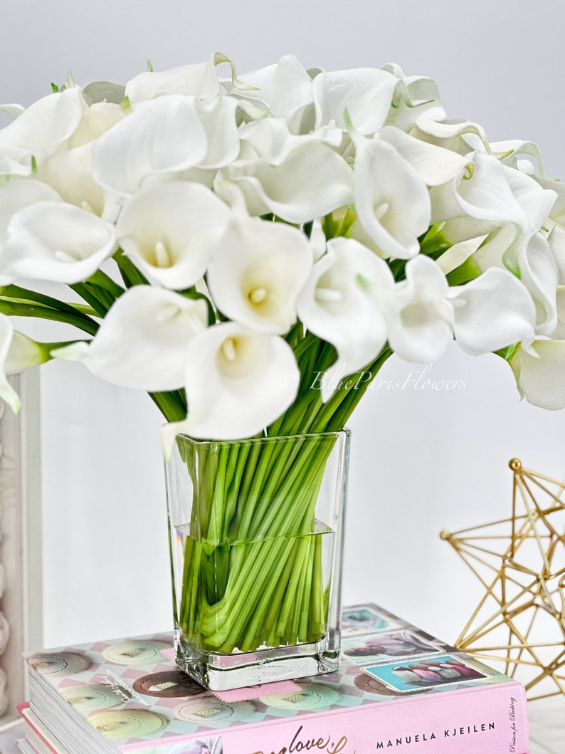 May include: A bouquet of white calla lilies in a clear glass vase. The vase is sitting on a stack of books with a gold star ornament in the background.