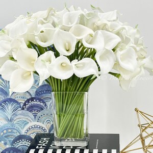May include: A bouquet of white calla lilies arranged in a clear glass vase. The vase is sitting on a black and white striped surface.