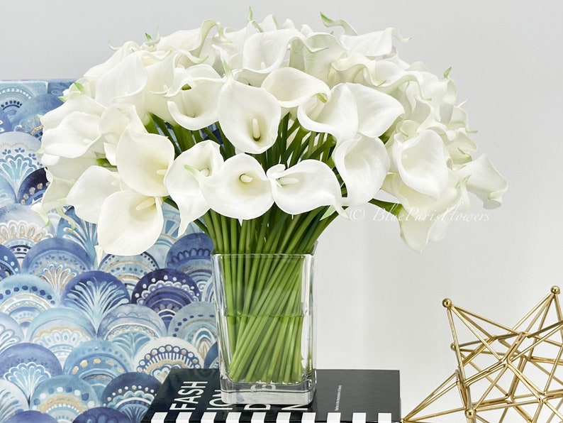 May include: A bouquet of white calla lilies arranged in a clear glass vase. The vase is sitting on a black and white striped surface.