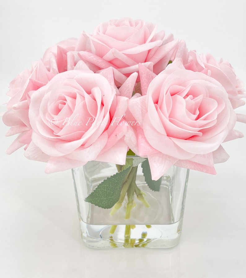 May include: A bouquet of pink silk roses arranged in a clear glass vase. The roses are in full bloom and have a soft, delicate appearance.