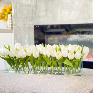May include: A glass vase filled with white tulips, arranged in a rectangular shape. The vase is sitting on a gray surface with a string of white lights in front of it.