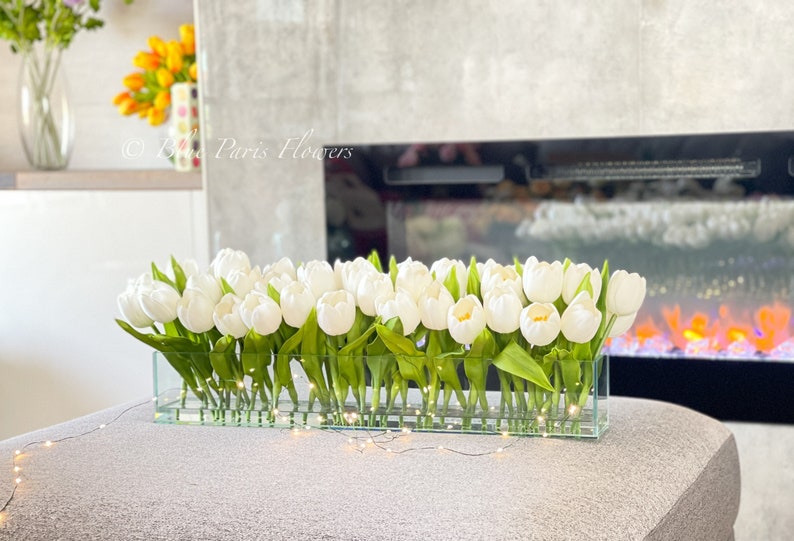 May include: A glass vase filled with white tulips, arranged in a rectangular shape. The vase is sitting on a gray surface with a string of white lights in front of it.