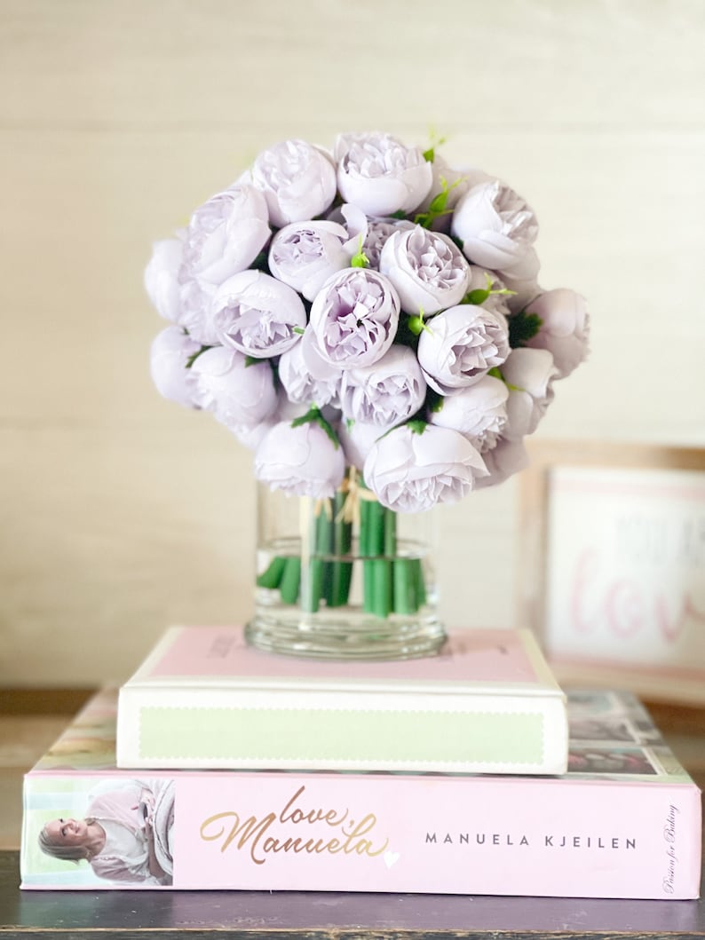 Pu&ograve; includere: Un bouquet di peonie artificiali color lavanda pallido in un vaso di vetro trasparente su due libri. Il libro superiore &egrave; rosa con una spina dorsale verde e il libro inferiore &egrave; rosa con il titolo "Love Manuela" e l'autore "MANUELA KJEILEN".
