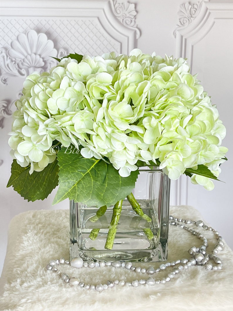 May include: A bouquet of light green hydrangeas in a clear glass vase. The vase is sitting on a white fluffy surface with a pearl necklace.
