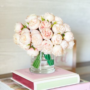 May include: A bouquet of pale pink peonies in a clear glass vase, sitting on top of two books. The top book has a pink cover with the text "love Manuela" in gold script. The bottom book has a white cover with the text "MANUELA KJEILEN" in black.