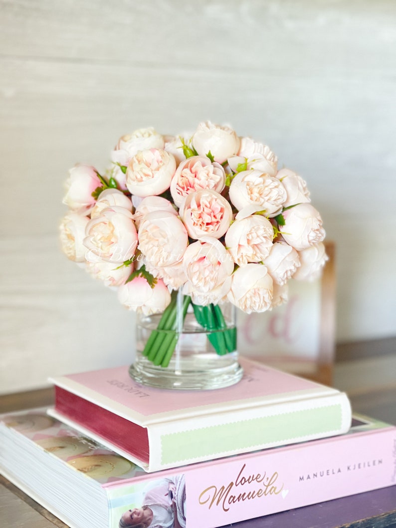 May include: A bouquet of pale pink peonies in a clear glass vase, sitting on top of two books. The top book has a pink cover with the text "love Manuela" in gold script. The bottom book has a white cover with the text "MANUELA KJEILEN" in black.