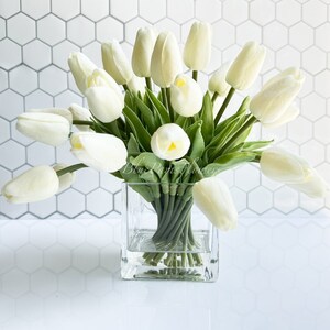 May include: A bouquet of white artificial tulips arranged in a clear glass cube vase. The vase is sitting on a white tiled surface.