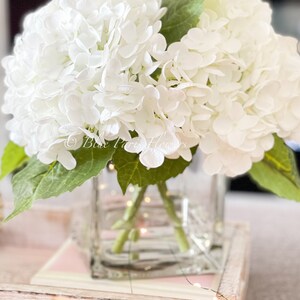 May include: A bouquet of white hydrangea flowers in a clear square glass vase. The flowers have green stems and leaves. The vase sits on a wooden tray with a pink and white design. The text "Blue Paris Flowers" is visible.