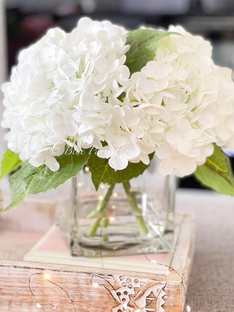 May include: A bouquet of white hydrangea flowers in a clear square glass vase. The flowers have green stems and leaves. The vase sits on a wooden tray with a pink and white design. The text "Blue Paris Flowers" is visible.