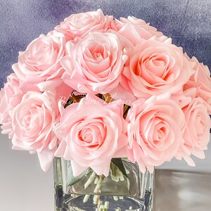 May include: A bouquet of pink silk roses arranged in a clear glass cube vase. The roses are in full bloom and have a soft, delicate appearance.