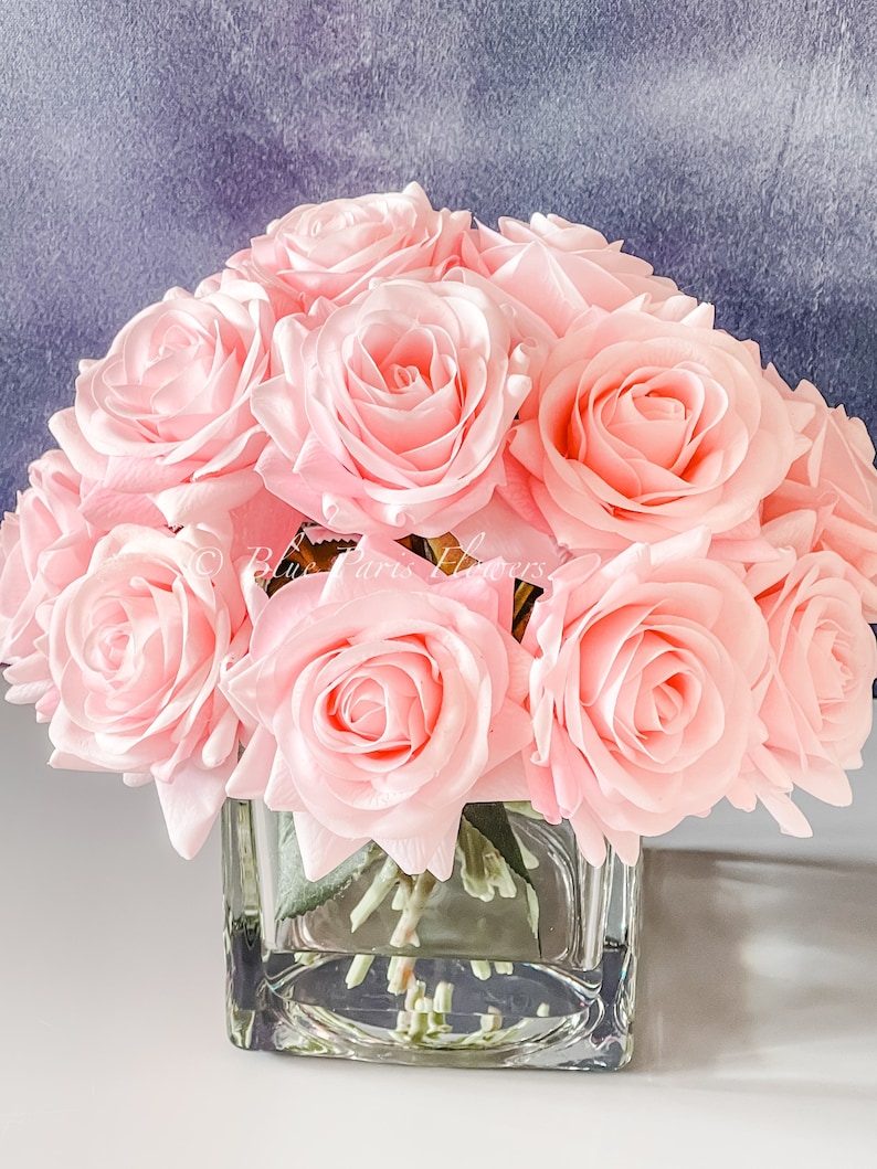May include: A bouquet of pink silk roses arranged in a clear glass cube vase. The roses are in full bloom and have a soft, delicate appearance.