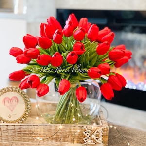 May include: A bouquet of red tulips in a clear glass vase, sitting on a wooden tray with a string of white lights. The vase is on a table with a small framed picture of a heart.