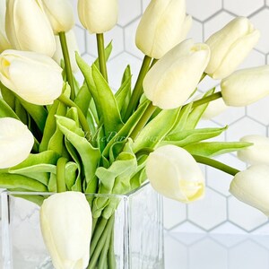 May include: A bouquet of white tulips arranged in a clear glass cube vase. The tulips are in full bloom and have green stems and leaves.