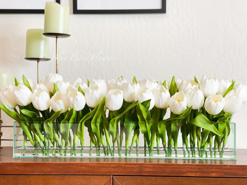 May include: A glass vase filled with white tulips. The vase is clear and rectangular. The tulips are arranged in a row and have green leaves.