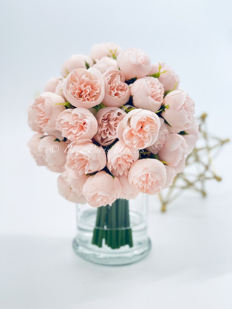 May include: A bouquet of blush pink silk peonies arranged in a clear glass vase. The vase has a textured base.