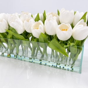 May include: A glass vase filled with white artificial tulips. The vase is clear and has a textured surface. The tulips are arranged in a row and have green stems.