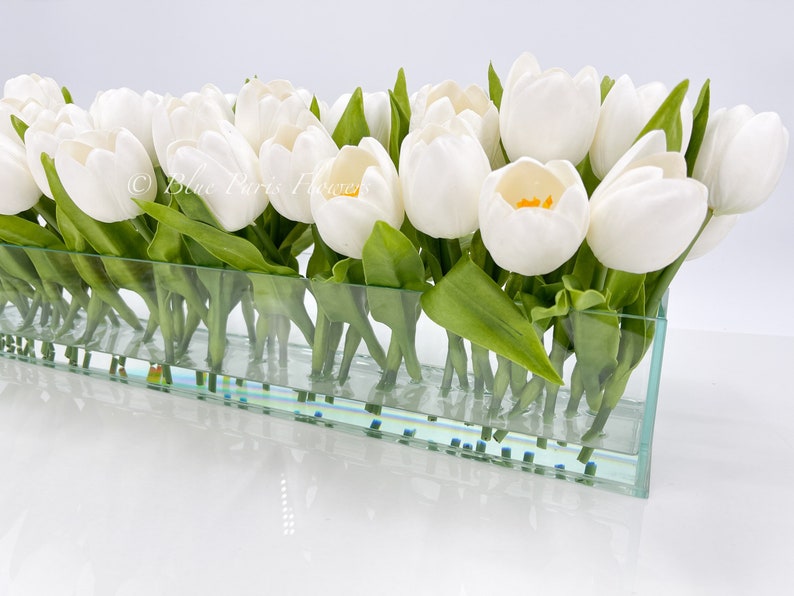 May include: A glass vase filled with white artificial tulips. The vase is clear and has a textured surface. The tulips are arranged in a row and have green stems.