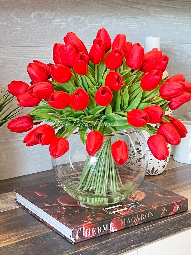 May include: A bouquet of red tulips in a clear glass vase. The vase is sitting on a book with the word "HERM&Egrave;S" printed on the cover.