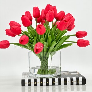 May include: A bouquet of red tulips in a clear glass vase. The vase is sitting on a black and white striped book with the text "Elements of Style" on the cover.