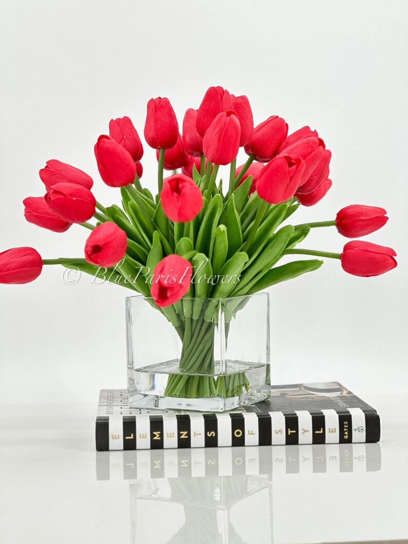May include: A bouquet of red tulips in a clear glass vase. The vase is sitting on a black and white striped book with the text "Elements of Style" on the cover.