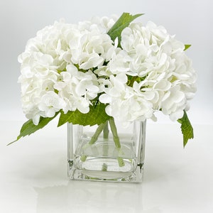 May include: A square glass vase filled with white hydrangea flowers and green leaves. The flowers are clustered together, creating a full, lush appearance. The vase sits on a white surface.