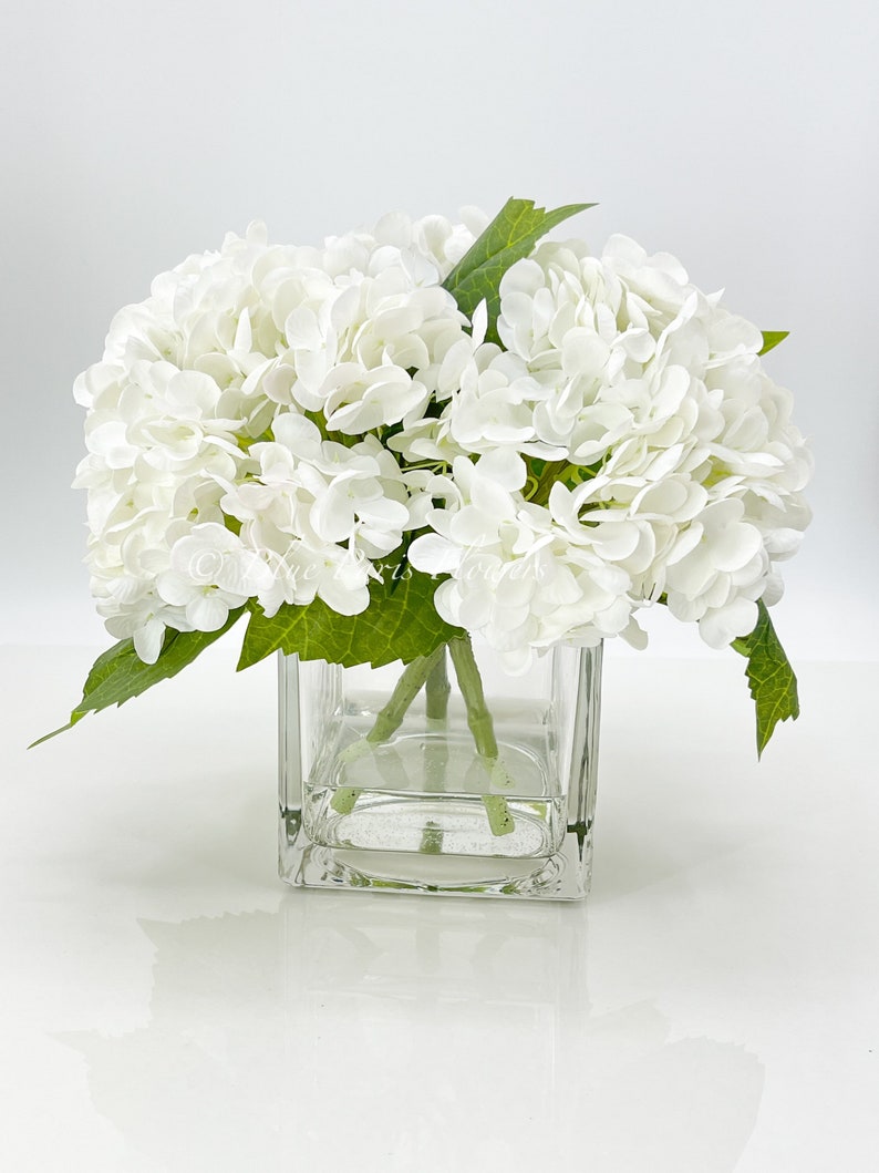 May include: A square glass vase filled with white hydrangea flowers and green leaves. The flowers are clustered together, creating a full, lush appearance. The vase sits on a white surface.