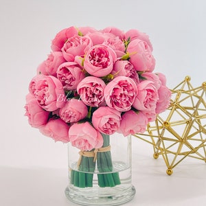 May include: A bouquet of pink peonies arranged in a clear glass vase. The vase is tied with a brown ribbon and the stems are green.