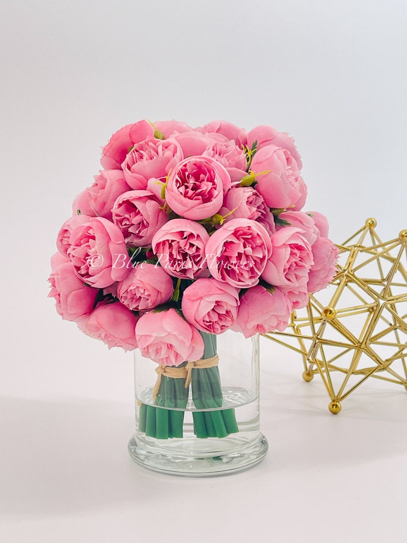 May include: A bouquet of pink peonies arranged in a clear glass vase. The vase is tied with a brown ribbon and the stems are green.