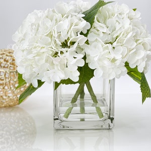 May include: A square glass vase holds a bouquet of white hydrangea flowers with green stems and leaves. A gold decorative sphere sits to the left of the vase. The arrangement is on a white surface.