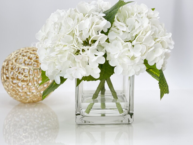 May include: A square glass vase holds a bouquet of white hydrangea flowers with green stems and leaves. A gold decorative sphere sits to the left of the vase. The arrangement is on a white surface.