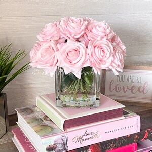 May include: A bouquet of pink silk roses in a clear glass cube vase. The vase is sitting on a stack of books with pink covers. The top book has the text "You are so loved" on the cover.