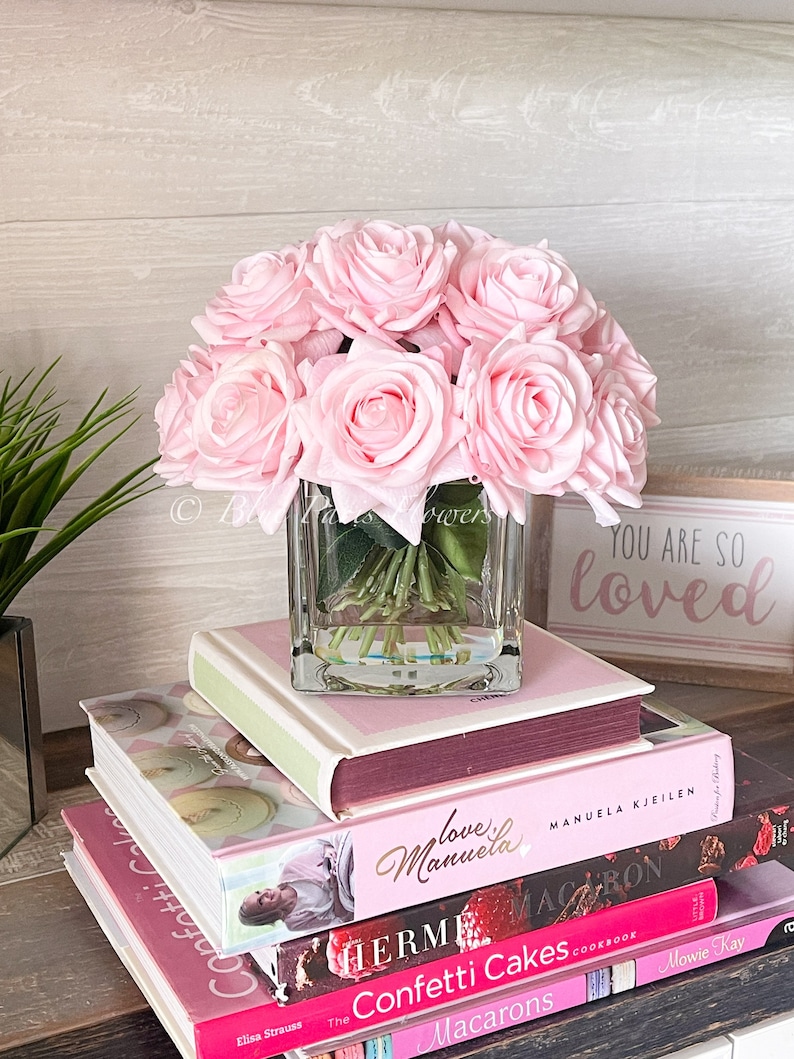 May include: A bouquet of pink silk roses in a clear glass cube vase. The vase is sitting on a stack of books with pink covers. The top book has the text "You are so loved" on the cover.