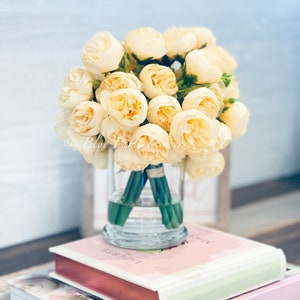 May include: A bouquet of cream-coloured silk peonies arranged in a clear glass vase. The vase is sitting on top of two books, one with the title "Love Manuela" and the other with the title "La Torte Rose".