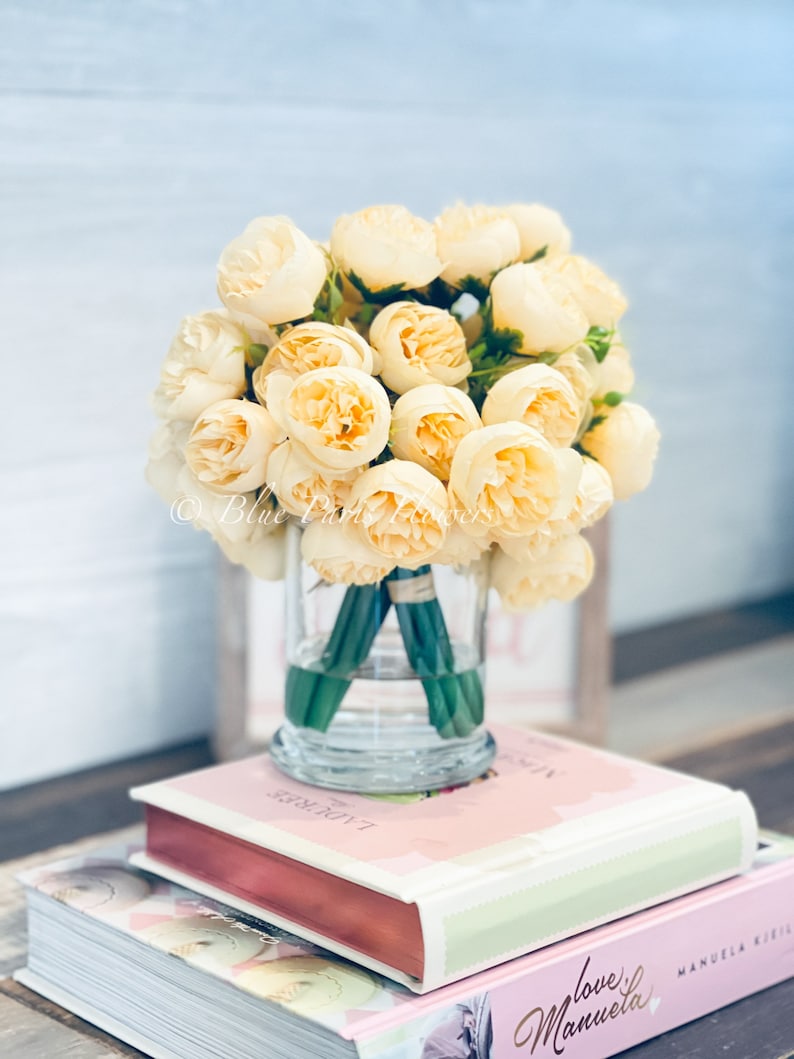 May include: A bouquet of cream-coloured silk peonies arranged in a clear glass vase. The vase is sitting on top of two books, one with the title "Love Manuela" and the other with the title "La Torte Rose".