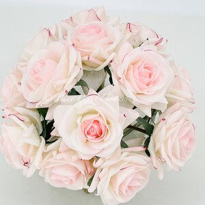 May include: A bouquet of 12 artificial pink roses with white centers and burgundy edges. The roses are arranged in a circular shape and are in a clear vase.