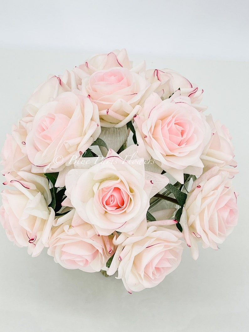 May include: A bouquet of 12 artificial pink roses with white centers and burgundy edges. The roses are arranged in a circular shape and are in a clear vase.