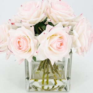 May include: A bouquet of pale pink roses with red edges on the petals, arranged in a clear glass cube vase.