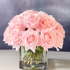 May include: A bouquet of pink silk roses arranged in a clear glass cube vase. The roses are in full bloom and have a realistic appearance.