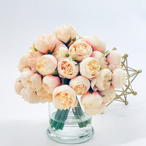 May include: A bouquet of blush pink artificial peonies arranged in a clear glass vase. The vase has a thick base and is sitting on a white surface.